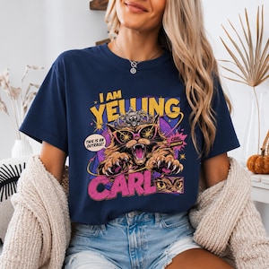 Puede incluir: Una camiseta azul marino con el gráfico de un gato que lleva una corona y gafas de sol rosas. El texto dice I AM YELLING, THIS IS AN OUTRAGE!, CARL y CARL, HONESTLY. El gato tiene garras afiladas y una expresión feroz sobre un fondo morado.