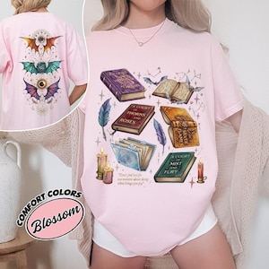 Könnte beinhalten: Hellrosa T-Shirt mit einem Fantasy-Buch-Design, das Bücher, Kerzen, Federn und Fledermäuse zeigt. Auf der Rückseite des Shirts befindet sich ein geflügeltes Augendesign. Das Shirt ist mit "Comfort Colors" und "Blossom" gekennzeichnet.