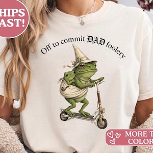 Könnte beinhalten: Cremefarbenes T-Shirt mit einer skurrilen Illustration eines grünen Frosches auf einem Roller, der einen Zaubererhut trägt und einen anderen Frosch trägt. Der Text lautet "Off to commit DAD foolery."