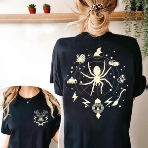 Könnte beinhalten: Schwarze T-Shirts mit cremefarbenen grafischen Designs. Ein Shirt zeigt einen Hirschkopf mit Blitzen und einer Fliege. Das andere Shirt hat ein Spinnennetz-Design mit verschiedenen Symbolen.