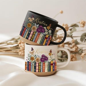 Könnte beinhalten: Zwei Keramikbecher, einer schwarz und einer cremefarben, gestapelt. Jeder Becher hat ein Blumen- und Buchdesign mit dem Namen "Kristem" in goldener Schrift. Die Becher haben einen naturbelassenen Boden und schwarze oder cremefarbene Griffe.