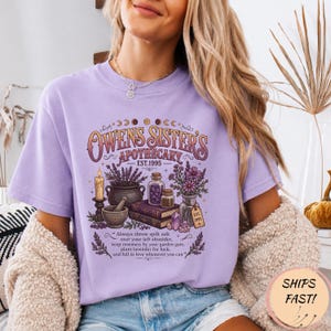 Può includere: Una t-shirt color viola chiaro con una stampa grafica che riporta il testo "Owens Sister's Apothecary Est. 1995" e "Always throw spilt salt over your left shoulder, keep rosemary by your garden gate, plant lavender for luck, and fall in love whenever you can."
