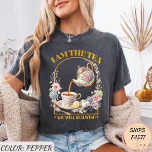 Può includere: T-shirt grigio scuro con grafica floreale a tema tè. Il design presenta una teiera che versa tè in una tazza, circondata da fiori, con la scritta "I AM THE TEA" e "YOU WILL BE HAVING".