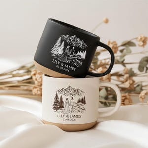 Può includere: Due tazze in ceramica, una nero opaco e una bianco sporco maculato, ciascuna con un'illustrazione a linee di una coppia davanti a montagne e pini. Il testo LILY & JAMES 03.08.2026 è stampato sotto l'illustrazione su entrambe le tazze.