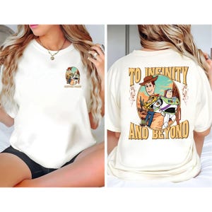 Op de afbeelding: Crèmekleurig T-shirt met een cartoonafbeelding van Woody en Buzz Lightyear. De voorkant heeft een kleine afbeelding met de tekst "CUSTOM NAME". De achterkant van het shirt heeft de tekst "TO INFINITY AND BEYOND" met een woestijnscène.