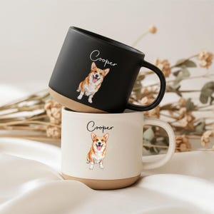 Könnte beinhalten: Zwei Keramikbecher, einer in mattem Schwarz und einer in gesprenkeltem Off-White, jeweils mit einer handgezeichneten Illustration eines Corgi-Hundes und dem Namen Cooper in Schreibschrift darüber. Beide Becher haben einen glatten, unglasierten beigen Boden.