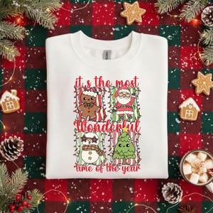 Könnte beinhalten: Weißes Sweatshirt mit einem festlichen Design mit einem Lebkuchenmann, dem Weihnachtsmann, einem Schneemann und einem Weihnachtsbaum. Der Text "it's the most wonderful time of the year" ist in Rot und Grün gedruckt.