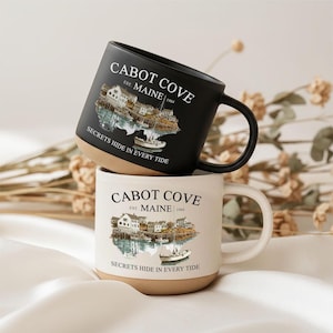 Puede incluir: Dos tazas de cerámica, una negra mate y otra de color blanco roto moteado, con una ilustración costera de Cabot Cove, Maine, establecida en 1984. El texto dice Cabot Cove, Est. Maine 1984, y Secrets Hide In Every Tide, con un gráfico de barco.