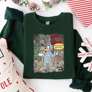 Könnte beinhalten: Dunkelgrünes Sweatshirt mit einem Cartoon von Goofy in Lametta, mit dem Text "Goofy's Tinsel Trouble!" und "Hyuck! That ain't how ya deck the halls!" Weihnachts-Design mit dekoriertem Baum und Geschenken.