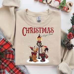 Könnte beinhalten: Beiges Sweatshirt mit dem Text "CHRISTMAS on Main Street" in Rot und Grün. Enthält eine Cartoon-Illustration von Mickey Mouse und Pluto in Winterkleidung, die neben einer Straßenlaterne stehen. Ein festliches Weihnachtsdesign.