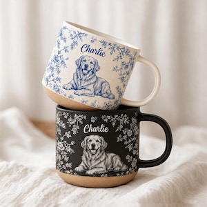 Könnte beinhalten: Zwei vertikal gestapelte Keramiktassen. Die obere Tasse ist cremefarben mit blauen Blumenmustern und einer blauen Illustration eines Golden Retrievers mit dem Namen Charlie. Die untere Tasse ist schwarz mit weißen Blumenmustern.
