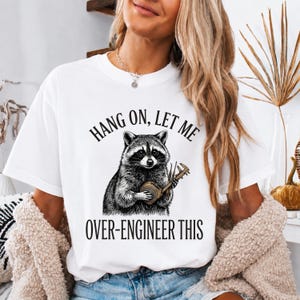 Könnte beinhalten: Ein weißes Kurzarm-T-Shirt mit einer schwarzen Grafik eines Waschbären, der einen Messschieber hält. Der Text über dem Waschbären lautet HANG ON, LET ME und der Text darunter lautet OVER-ENGINEER THIS in einer fetten, schwarzen Schriftart.