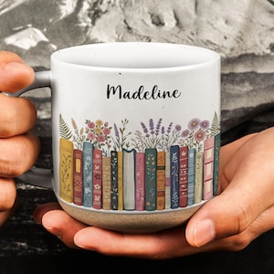 Könnte beinhalten: Ein weißer Keramikbecher mit gesprenkelter Textur am Boden, bedruckt mit einer Reihe bunter Bücher, aus denen Wildblumen wachsen. Der Name Madeline ist in schwarzer Schreibschrift über den illustrierten Buchrücken aufgedruckt.