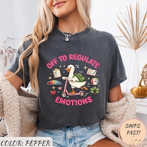 Könnte beinhalten: Dunkelgraues T-Shirt mit dem Text "OFF TO REGULATE EMOTIONS". Die Grafik zeigt eine Cartoon-Ente auf einem rosa Roller, mit einem Rucksack und verschiedenen bunten Symbolen. Das Shirt hat die Farbe Pfeffer.