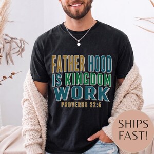 Könnte beinhalten: Schwarzes T-Shirt mit dem Text "FATHER HOOD IS KINGDOM WORK" in Gold, Grün und Türkis. Der Text "PROVERBS 22:6" steht darunter. Das Shirt ist dunkel und hat einen Vintage-Look.