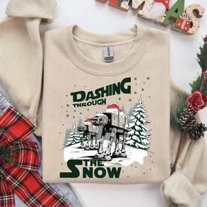 Op de afbeelding: Beige sweatshirt met een afbeelding van AT-AT's met kerstmutsen in een besneeuwd landschap met kerstbomen. De tekst luidt "Dashing Through The Snow" in groen en wit.