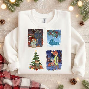 Puede incluir: Sudadera blanca con cuatro ilustraciones cuadradas de personajes de dibujos animados en escenas invernales. Las ilustraciones incluyen un perezoso y un búfalo, una serpiente, un castor con un árbol de Navidad y un zorro y un conejo. Decorada con ramas de pino y piñas.