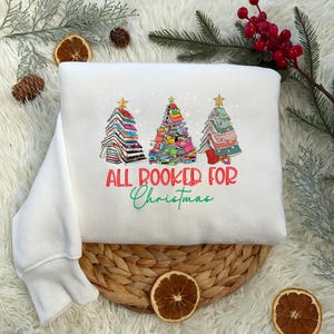 Könnte beinhalten: Weißes Sweatshirt mit einer Grafik von drei Weihnachtsbäumen aus Büchern. Der Text "ALL BOOKED FOR Christmas" ist darunter in Rot und Grün gedruckt. Das Sweatshirt wird auf einer gewebten Matte präsentiert.