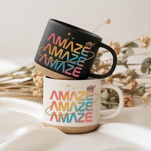 Könnte beinhalten: Zwei Keramikbecher, einer schwarz und einer cremefarben, gestapelt. Jeder Becher zeigt das Wort "AMAZE" in einem Regenbogenverlauf, mit einer Kamelillustration und einem goldenen Ringdesign. Die Becher haben einen beigefarbenen Boden und schwarze Griffe.