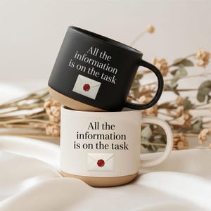 Könnte beinhalten: Zwei Keramikbecher, einer schwarz und einer weiß, übereinander gestapelt. Jeder Becher hat einen beigen Boden und einen schwarzen Henkel. Der schwarze Becher hat den Text "All the information is on the task" und eine weiße Umschlag-Grafik. Der weiße Becher hat den gleichen Text und die gleiche Grafik.