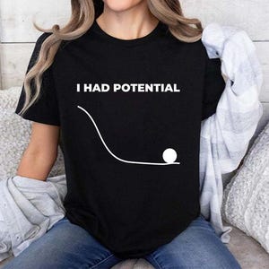 Pode incluir: Camiseta preta com o texto branco "I HAD POTENTIAL" acima de um gráfico branco de uma bola rolando por um caminho curvo. A camisa é de manga curta.