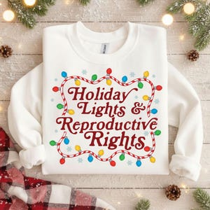 Peut inclure: Un sweat-shirt blanc avec l'inscription "Holiday Lights & Reproductive Rights" dans un design festif. Le texte est encadré par une guirlande de lumières de Noël colorées. Le sweat-shirt est de style col rond.