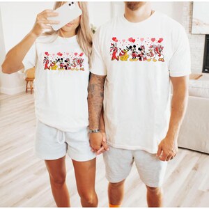 Pu&ograve; includere: Due magliette bianche con una grafica di personaggi Disney che tengono palloncini a forma di cuore. I personaggi includono Topolino, Minnie, Paperino, Paperina e Pippo. Le magliette sono indossate da due persone.