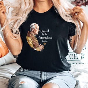 Könnte beinhalten: Schwarzes T-Shirt mit einer Illustration einer religiösen Figur in goldenen und roten Gewändern. Der Text "Blessed be the Peacemakers" ist in Weiß gedruckt, zusammen mit dem Namen "Peoleo".
