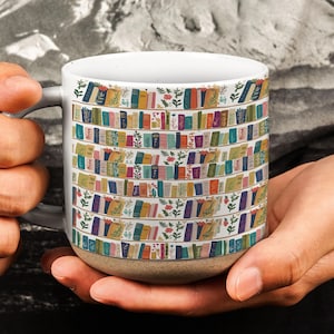 Könnte beinhalten: Ein weißer Keramikbecher mit gesprenkeltem beigefarbenem Boden, der ein sich wiederholendes Muster aus bunten Büchern in Regalen zeigt, durchsetzt mit kleinen grünen Blattzweigen und winzigen roten Blumen auf der gesamten Außenfläche.