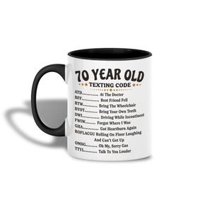 Könnte beinhalten: Eine weiße Keramiktasse mit schwarzem Innenraum und Griff. Die Tasse zeigt den Text "70 YEAR OLD TEXTING CODE" mit einer Liste von Akronymen und ihren Bedeutungen, wie "ATD... At The Doctor" und "FWIW... Forgot Where I Was."