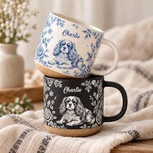 May include: Two ceramic mugs with a natural tan base. The top mug is white with blue floral patterns and a Cavalier King Charles Spaniel illustration with the name Charlie. The bottom mug is black with white floral patterns and the same dog.