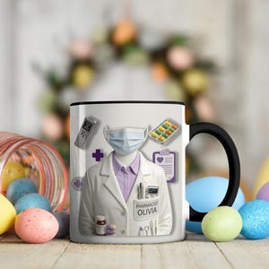 Könnte beinhalten: Eine weiße Keramik-Tasse mit schwarzem Henkel und dem Text "But First, Coffee" in fetter, schwarzer Schrift. Die Tasse steht vor einem verschwommenen Hintergrund, der eine gemütliche Umgebung suggeriert.
