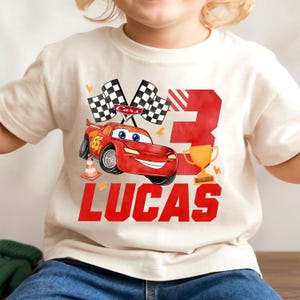 Könnte beinhalten: Cremefarbenes T-Shirt mit einem roten Cartoon-Rennwagen, der Nummer 95, karierten Flaggen und dem Namen "LUCAS" in Rot. Eine große rote Zahl 3 und eine goldene Trophäe sind ebenfalls vorhanden. Für Kinder.