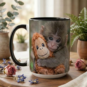 Puede incluir: Taza de cerámica blanca con asa y borde negros. La taza presenta una fotografía de un bebé mono abrazando a un mono de peluche. El mono de peluche es marrón y el bebé mono es gris.