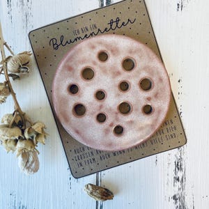Könnte beinhalten: Ein runder, gesprenkelter, rosa Keramik-Blumenfrosch mit mehreren Löchern, der auf einer braunen Karte mit deutschem Text ruht. Die Karte hat auch kleine schwarze Punkte und zusätzlichen Text.