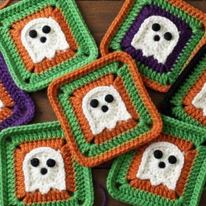 Pode incluir: Uma coleção de quadrados coloridos de crochê com tema de Halloween. Cada quadrado apresenta um design de fantasma branco com olhos de botão pretos, cercado por bordas laranja, verdes e roxas. Uma agulha de crochê e fios também são visíveis.