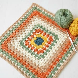 May include: A square, handmade crochet blanket in a granny square pattern. The blanket features a central blue square surrounded by yellow, orange, green, and cream-colored squares. Two balls of yarn, green and yellow, and a crochet hook are next to the blanket.
