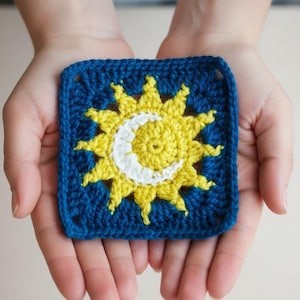 May include: A square, crocheted piece featuring a sun and moon design. The sun is yellow with pointed rays, and the moon is white. The border is a deep blue, creating a striking contrast. The piece is held in open hands.