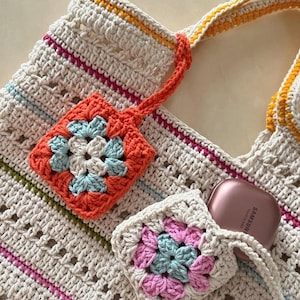 Op de afbeelding: Een cr&egrave;mekleurige gehaakte tote bag met oranje, roze en gele strepen. Twee vierkante gehaakte pouches met bloemmotieven in oranje, roze en blauw zijn bevestigd. Een roze Samsung-hoesje is zichtbaar in de tas.