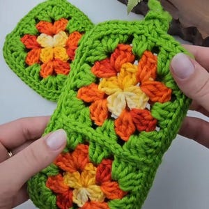 Op de afbeelding: Twee handgemaakte, groene gehaakte pannenlappen met een bloemenontwerp. De bloemen zijn gemaakt van oranje, gele en cr&egrave;mekleurige draad. Een pannenlap wordt door een persoon vastgehouden. De pannenlappen hebben een lus om op te hangen.