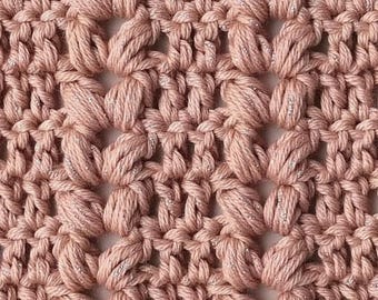 Puede incluir: Primer plano de una tela de ganchillo en un suave color rosa apagado. La textura presenta un patrón repetitivo de puntadas abultadas en relieve, intercaladas con espacios abiertos. El hilo tiene un sutil hilo metálico tejido, que añade un toque de brillo.