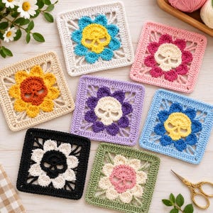 May include: Seven square crochet motifs featuring a central skull surrounded by flower petals in various colors including yellow, pink, purple, blue, white, and black, arranged on a light wooden surface with gold scissors nearby.