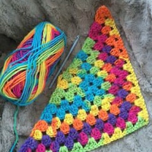 May include: A colorful crochet project in progress. A ball of rainbow yarn sits next to a partially completed granny square triangle in shades of purple, blue, green, yellow, and orange. A crochet hook is also visible.
