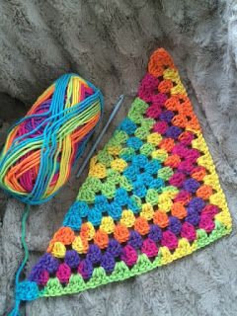 May include: A colorful crochet project in progress. A ball of rainbow yarn sits next to a partially completed granny square triangle in shades of purple, blue, green, yellow, and orange. A crochet hook is also visible.