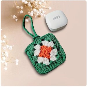 Op de afbeelding: Een handgemaakt groen, wit en oranje gehaakt vierkant met een groene handgreep. Een wit, afgerond vierkant object met drie kleine cirkels staat ook in de afbeelding. De items staan op een lichtroze ondergrond.