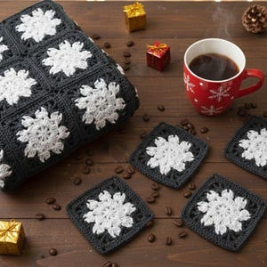 May include: A cozy scene featuring a black and white crocheted blanket and matching coasters with a snowflake design. A red mug of coffee, small gifts, pine cones, and coffee beans are arranged on a wooden surface, creating a warm, inviting atmosphere.