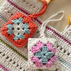 May include: Two crocheted square pouches with floral designs. One pouch is orange with a blue flower, the other is pink with a blue flower. Both pouches are attached to a crocheted bag with a white, orange, and yellow striped strap.