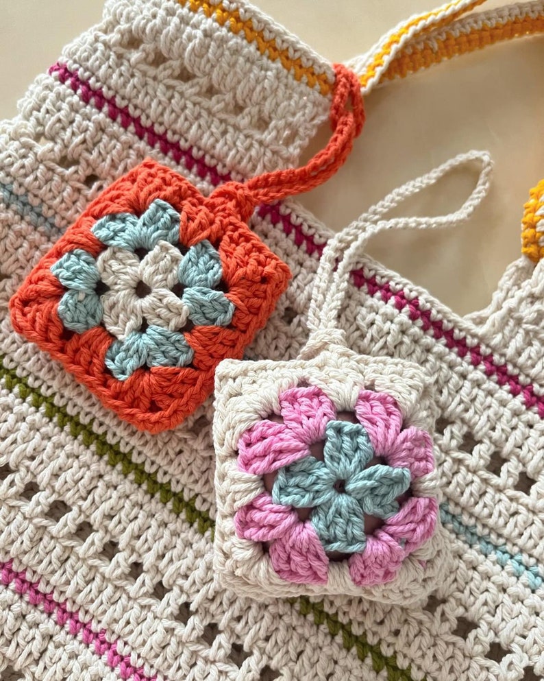 May include: Two crocheted square pouches with floral designs. One pouch is orange with a blue flower, the other is pink with a blue flower. Both pouches are attached to a crocheted bag with a white, orange, and yellow striped strap.
