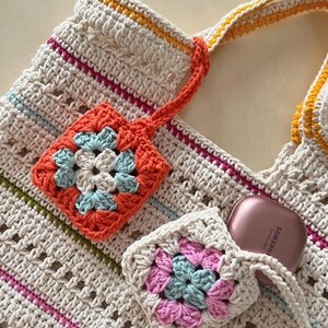 May include: A crocheted tote bag with a cream background and colorful stripes. The bag has two crocheted flower-shaped pockets, one orange and one pink, with a blue center. A small pink and blue crocheted pouch is attached to the bag, and a rose gold Samsung Galaxy Buds case is inside the pouch.