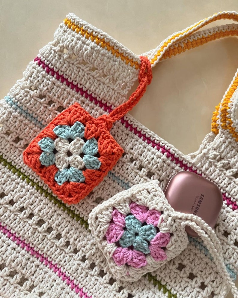 May include: A crocheted tote bag with a cream background and colorful stripes. The bag has two crocheted flower-shaped pockets, one orange and one pink, with a blue center. A small pink and blue crocheted pouch is attached to the bag, and a rose gold Samsung Galaxy Buds case is inside the pouch.